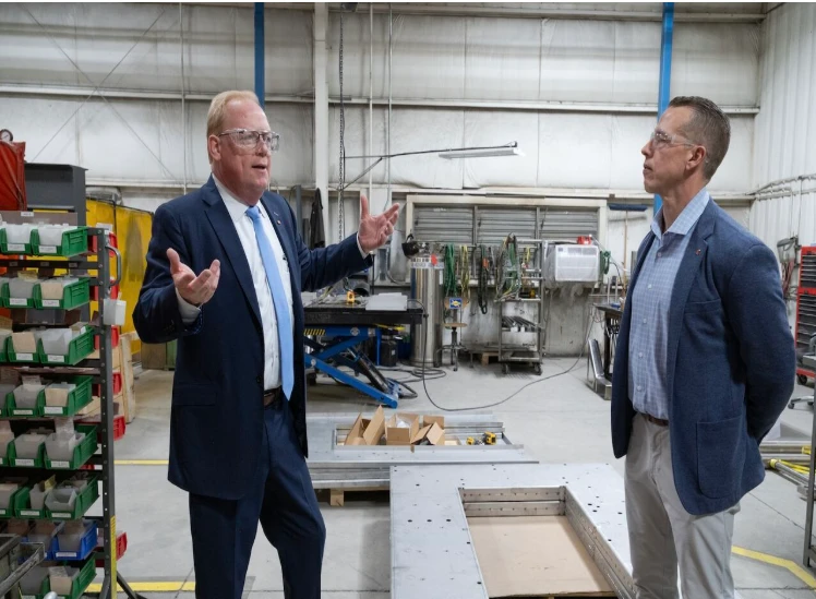 Two people stand and talk inside an industrial workshop. One, dressed in a dark blue suit and glasses, gestures with both hands while speaking. The other, wearing a blue blazer and light pants, listens with hands in pockets. Behind them are shelves with colored bins, metal machinery, and a large metallic frame on the floor, creating a busy manufacturing setting.