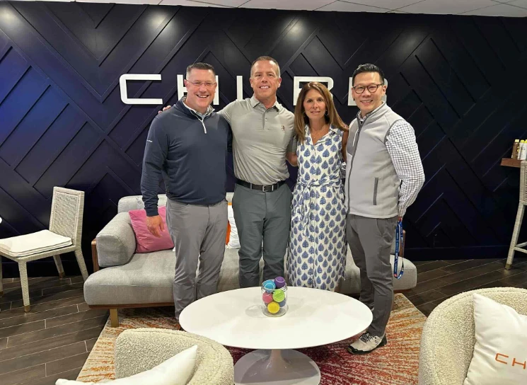 Four people stand together in a modern lounge area with a dark geometric wall behind them featuring large white letters spelling “CHUBB.” They wear business‑casual clothing, including golf shirts and a patterned dress. A gray couch with colorful pillows and a white round coffee table holding a small bowl of bright balls sit in front of them, creating a contemporary office setting.