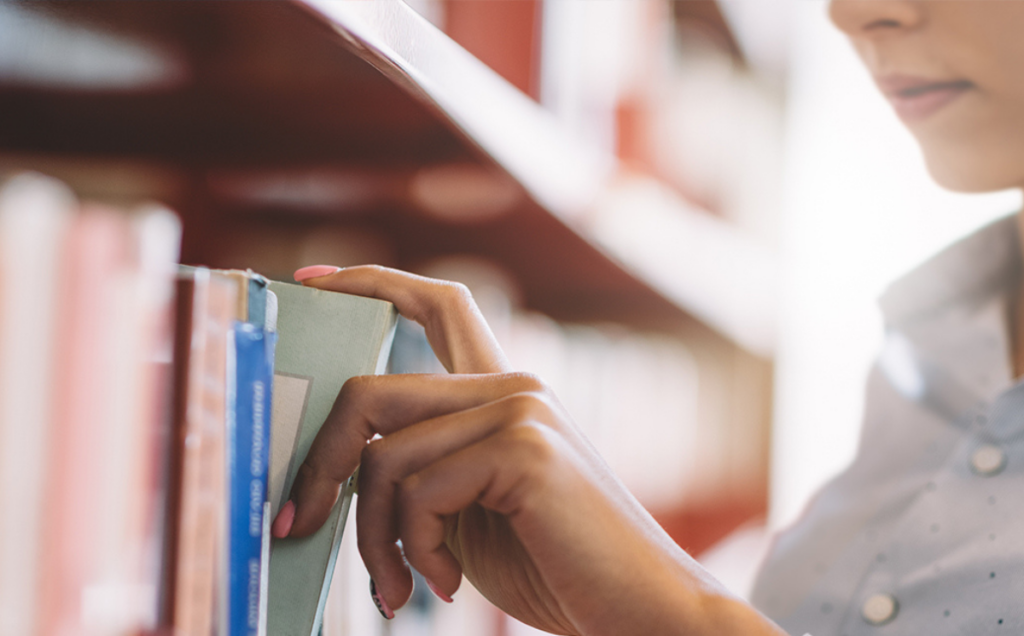 A hand pulls a light green book from a shelf lined with colorful spines in a quiet library or bookstore.