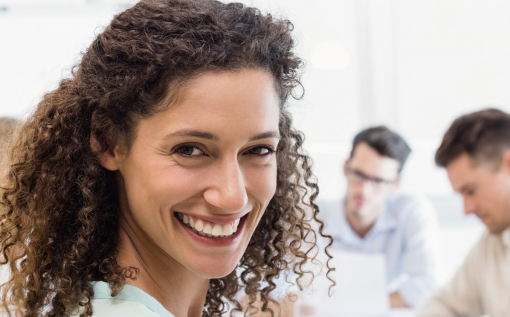 A person with curly hair smiles at the camera in the foreground while two people sit at a table in the background, appearing engaged in discussion in a bright, modern office setting.