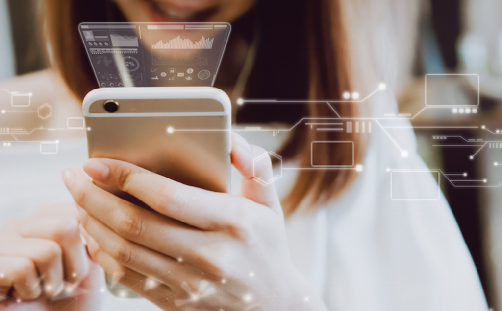 A person holds a smartphone with both hands while holographic-style digital graphics—charts, graphs, and network connections—float above the screen, suggesting advanced data visualization or futuristic mobile technology.