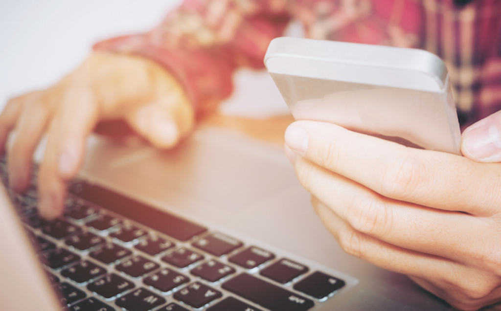 A person in a red plaid shirt types on a laptop with one hand while holding a smartphone in the other, illustrating multitasking across digital devices.