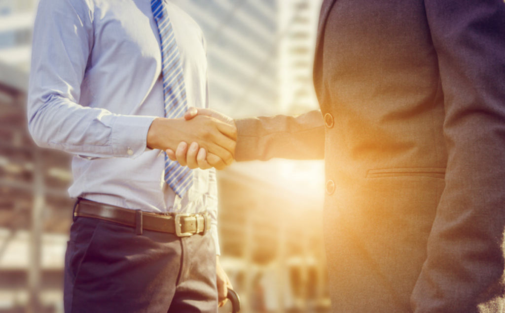 Two people in business attire shake hands outdoors with sunlight behind them, standing in front of modern glass buildings, symbolizing partnership or professional agreement.