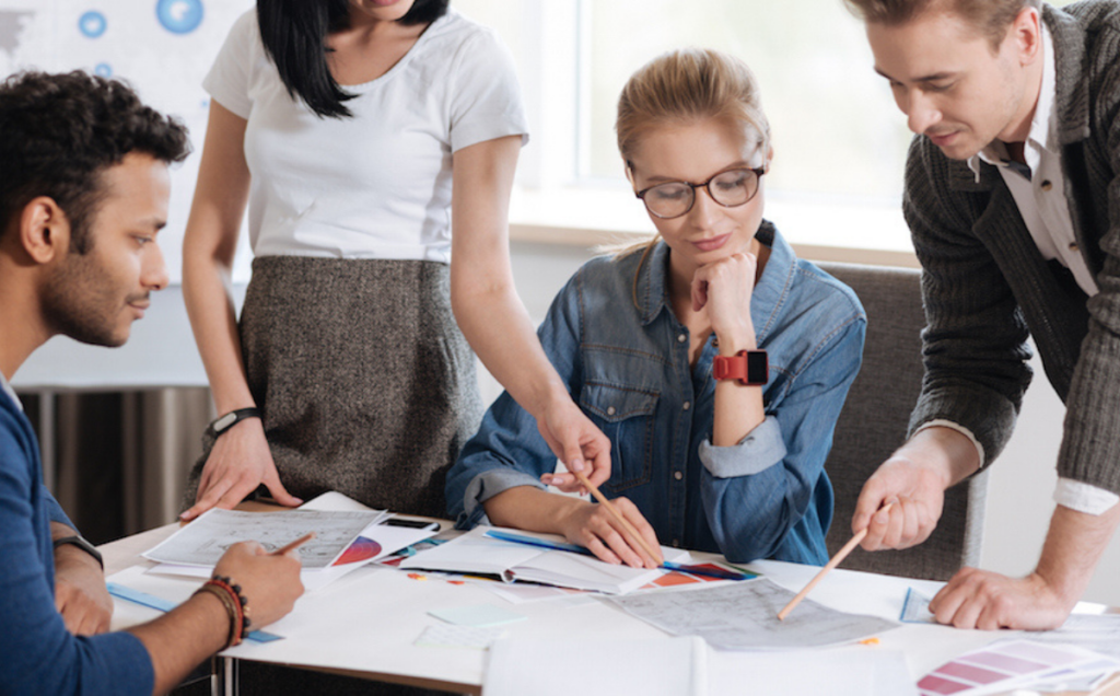 Four people gather around a table covered with papers and colorful charts, pointing and discussing ideas in a bright modern office, illustrating teamwork and collaborative planning.