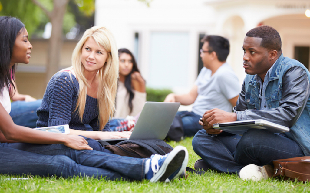 Students sit together on a grassy campus lawn, talking and working with notebooks, a laptop, and a tablet, with trees and an arched building in the background.