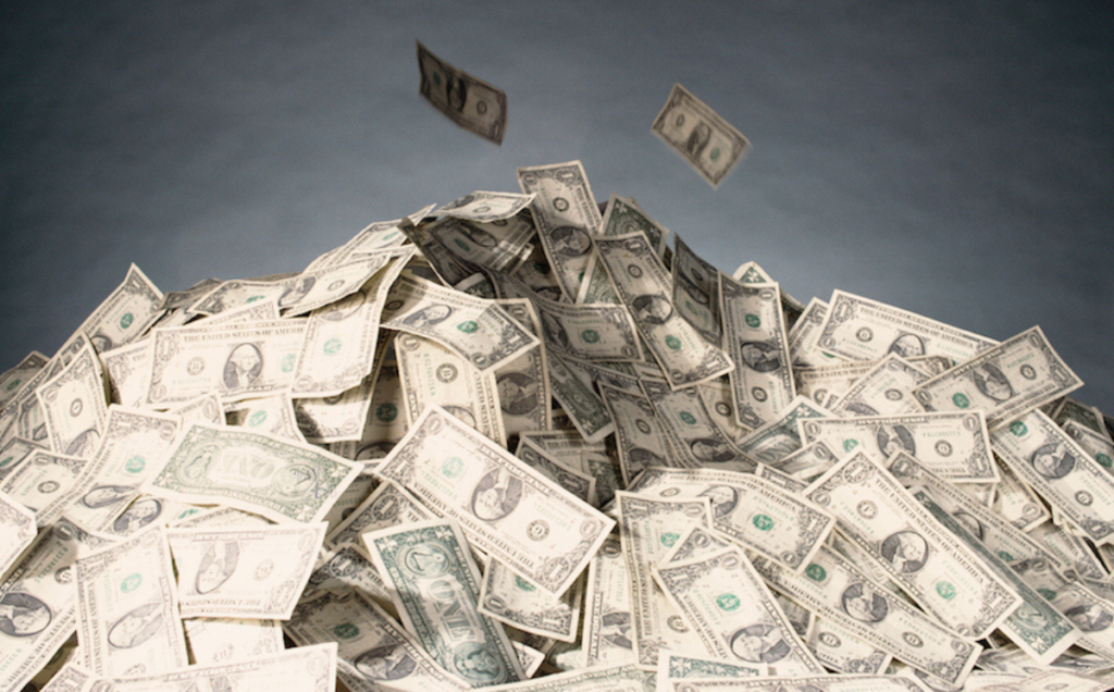 A large pile of U.S. one‑dollar bills sits in a heap, with a few bills appearing to float above it against a neutral gray background.