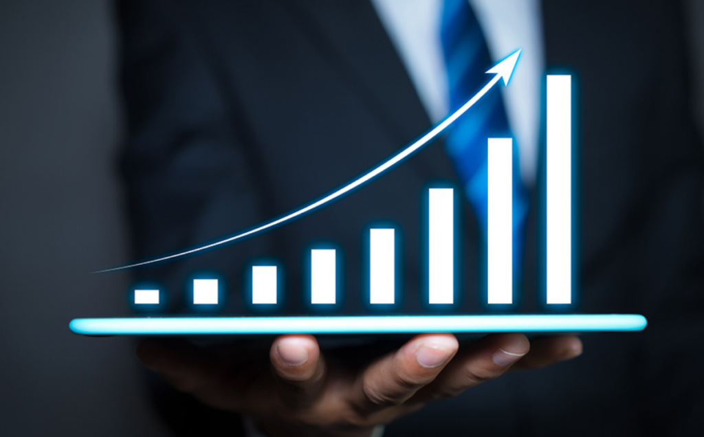 A person in a business suit holds a tablet projecting a glowing holographic bar chart with rising bars and an upward arrow, symbolizing growth and progress against a dark background.