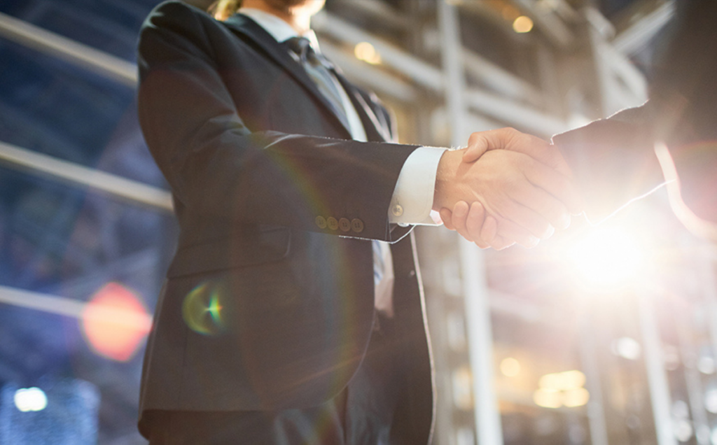 Two people in formal business attire shake hands in a bright modern building, with sunlight streaming in behind them, symbolizing partnership or professional agreement.