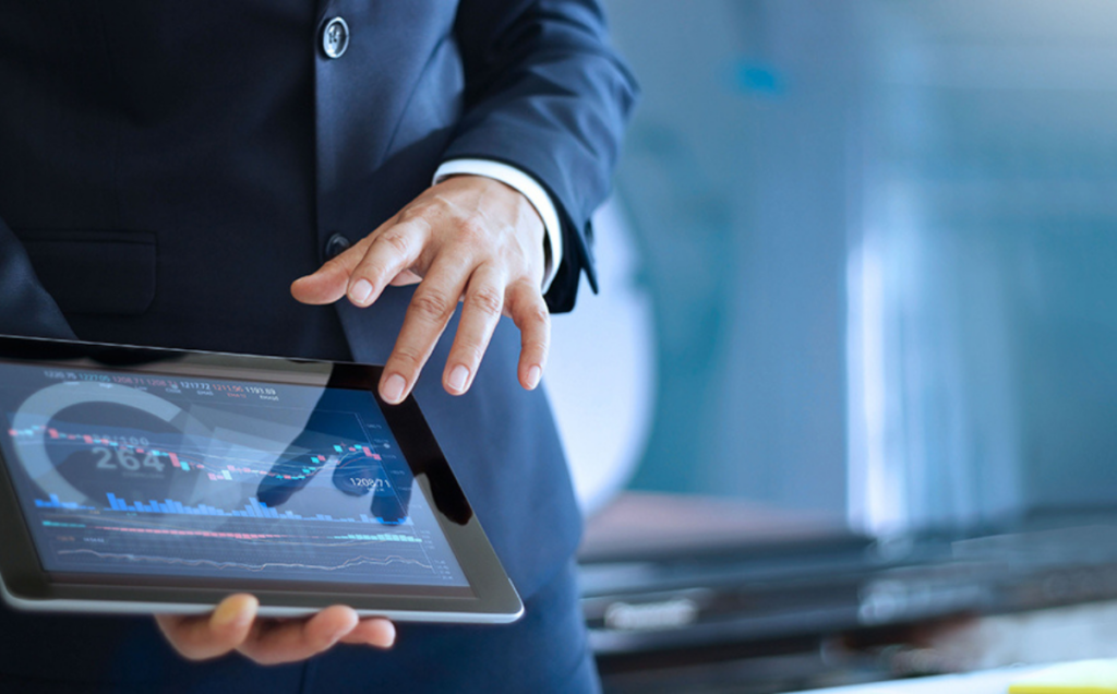 A person in a dark business suit holds a tablet displaying a colorful financial chart with lines, bars, and numerical data, suggesting stock market or trading analytics in a professional office setting.