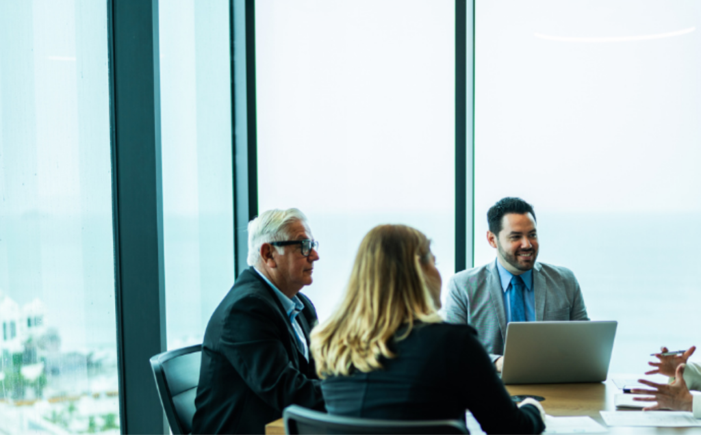 Four people sit around a conference table in a modern office with large glass windows overlooking the water. One person works on an open laptop while the others talk, creating a relaxed professional meeting atmosphere filled with natural light and a scenic view