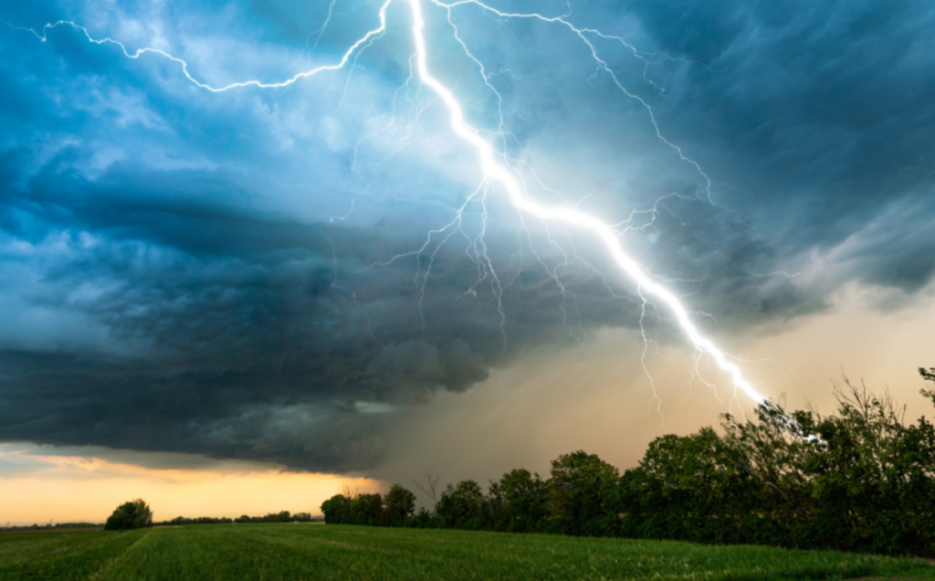 A bright lightning bolt strikes down from a dark, turbulent sky onto a landscape of green fields and trees. The clouds are heavy and stormy, with a band of lighter yellow sky near the horizon that contrasts sharply with the dramatic flash.