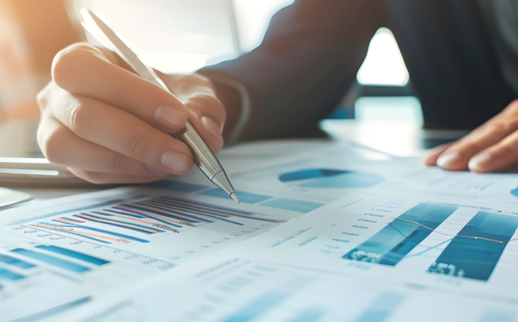 A close-up of a hand holding a pen while pointing to printed documents filled with blue-toned business charts, including bar graphs, pie charts, and line graphs, suggesting active data review or financial analysis in a professional setting.
