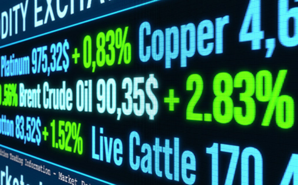 A digital board shows real‑time commodity prices with bright blue, green, and yellow highlights. Listed items include Platinum at 975.32 USD (+0.83%), Brent Crude Oil at 90.35 USD (+2.83%), Cotton at 83.52 USD (+1.52%), Copper at 4.6, and Live Cattle at 170.4, resembling an active trading interface.