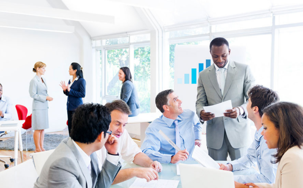A modern office scene shows several people collaborating. In the foreground, six individuals sit around a table covered with papers while one person stands, holding documents and speaking to the group. In the background, three others talk near large windows, and a presentation board displays a chart with rising blue bars. The space is bright, open, and professional, emphasizing teamwork and communication.