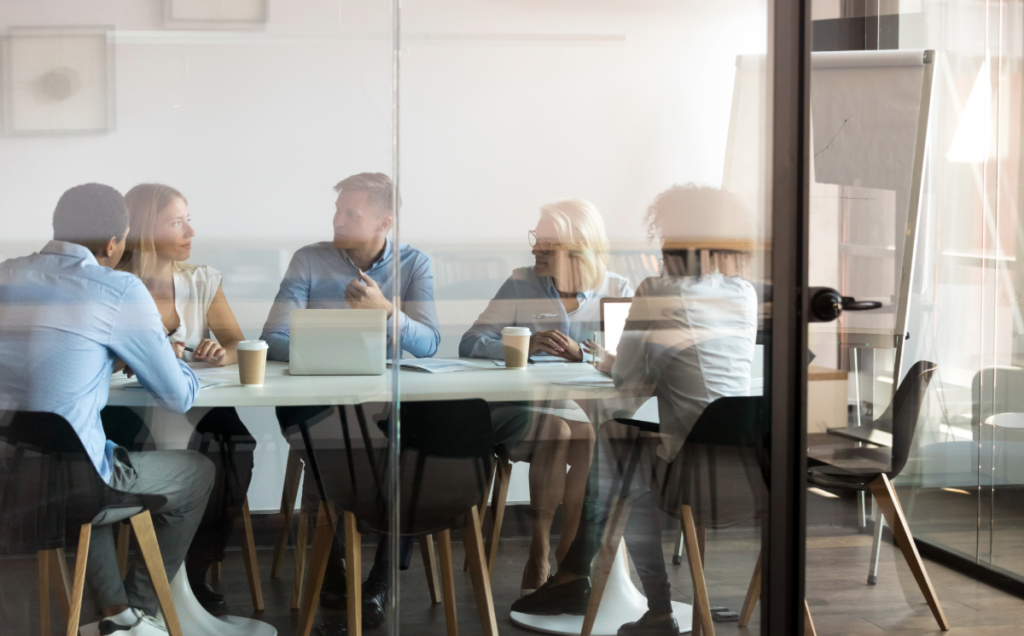 Five people sit around a conference table in a glass-walled meeting room, engaged in a business discussion. Laptops, notebooks, and takeaway coffee cups are on the table, while a flip chart stands in the background. The image is viewed through the glass wall, creating reflections that frame the collaborative workplace setting.