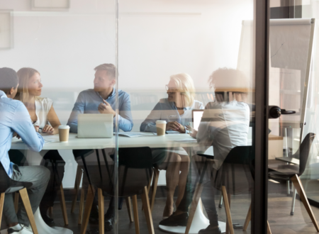 Five people sit around a conference table in a glass-walled meeting room, engaged in a business discussion. Laptops, notebooks, and takeaway coffee cups are on the table, while a flip chart stands in the background. The image is viewed through the glass wall, creating reflections that frame the collaborative workplace setting.